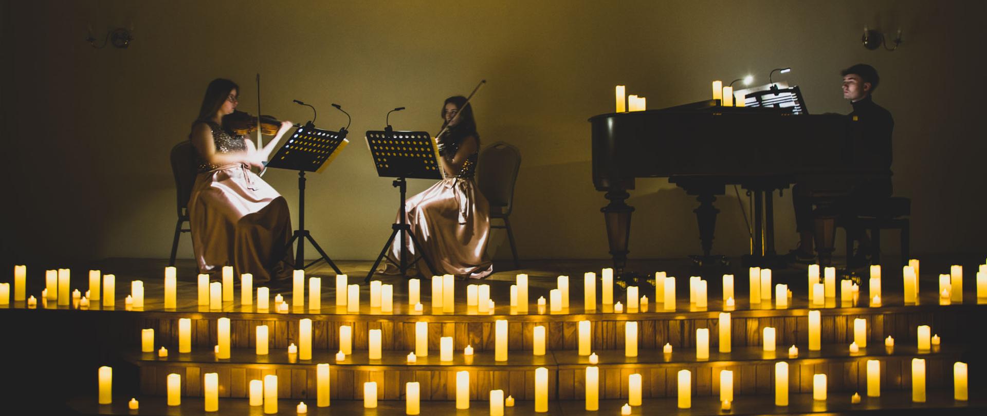 Skrzypaczka i altowiolistka grające na siedząco przy pulpitach oświetlonych lampkami. W pianista wykonujący swoją partię wy nutach oświetlonych lamką. Scena ozdobiona dużą ilością ozdobnych świec