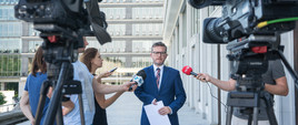 Minister Grzegorz Witkowski odpowiada na pytania dziennikarzy przed kamerami. W tle budynek ministerstwa i biurowiec.