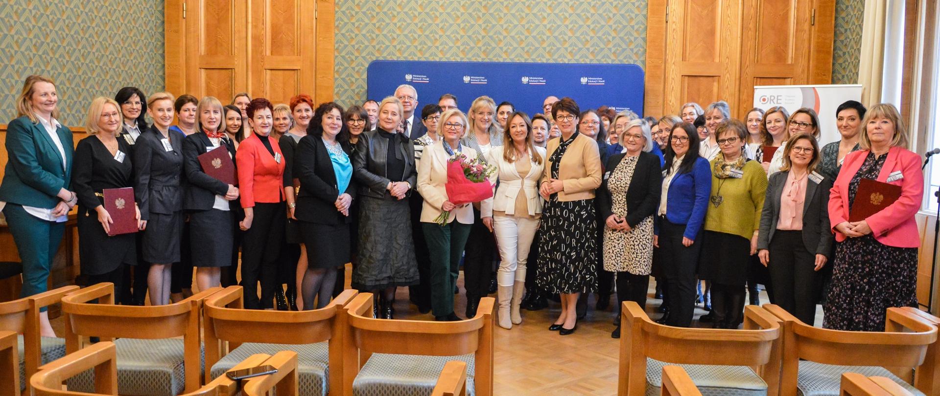 Zdjęcie zbiorowe, pod zieloną ścianą z dwojgiem drewnianych drzwi stoi duża grupa kolorowo ubranych ludzi, przed nimi puste drewniane krzesła ustawione w rzędy, po prawej okno.