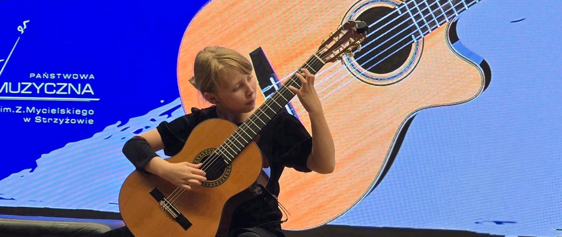 Na zdjęciu widoczna jest młoda dziewczyna, siedząca na krześle i grająca na gitarze klasycznej. Ubrana jest w czarną koszulkę i spodnie. Za nią znajduje się duży ekran, na którym wyświetlany jest wizerunek gitary oraz napisy: „XV MAKROREGIONALNE PRZESŁUCHANIA UCZNIÓW KLAS GITARY ZESPOŁÓW GITAROWYCH”. W górnej części ekranu widoczne są reflektory sceniczne.