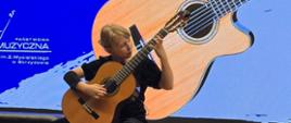 Na zdjęciu widoczna jest młoda dziewczyna, siedząca na krześle i grająca na gitarze klasycznej. Ubrana jest w czarną koszulkę i spodnie. Za nią znajduje się duży ekran, na którym wyświetlany jest wizerunek gitary oraz napisy: „XV MAKROREGIONALNE PRZESŁUCHANIA UCZNIÓW KLAS GITARY ZESPOŁÓW GITAROWYCH”. W górnej części ekranu widoczne są reflektory sceniczne.