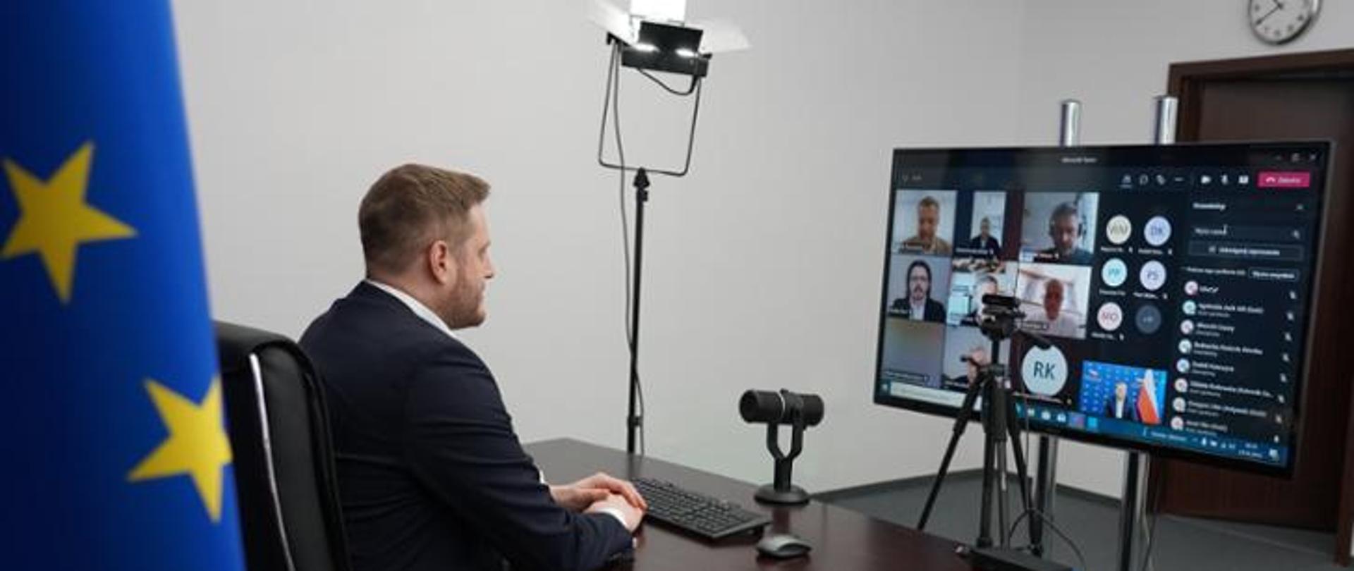 Minister Janusz Cieszyński siedzi przy drewnianym biurku przed nim monitor komputera podczas spotkania z dziennikarzami on-line. 