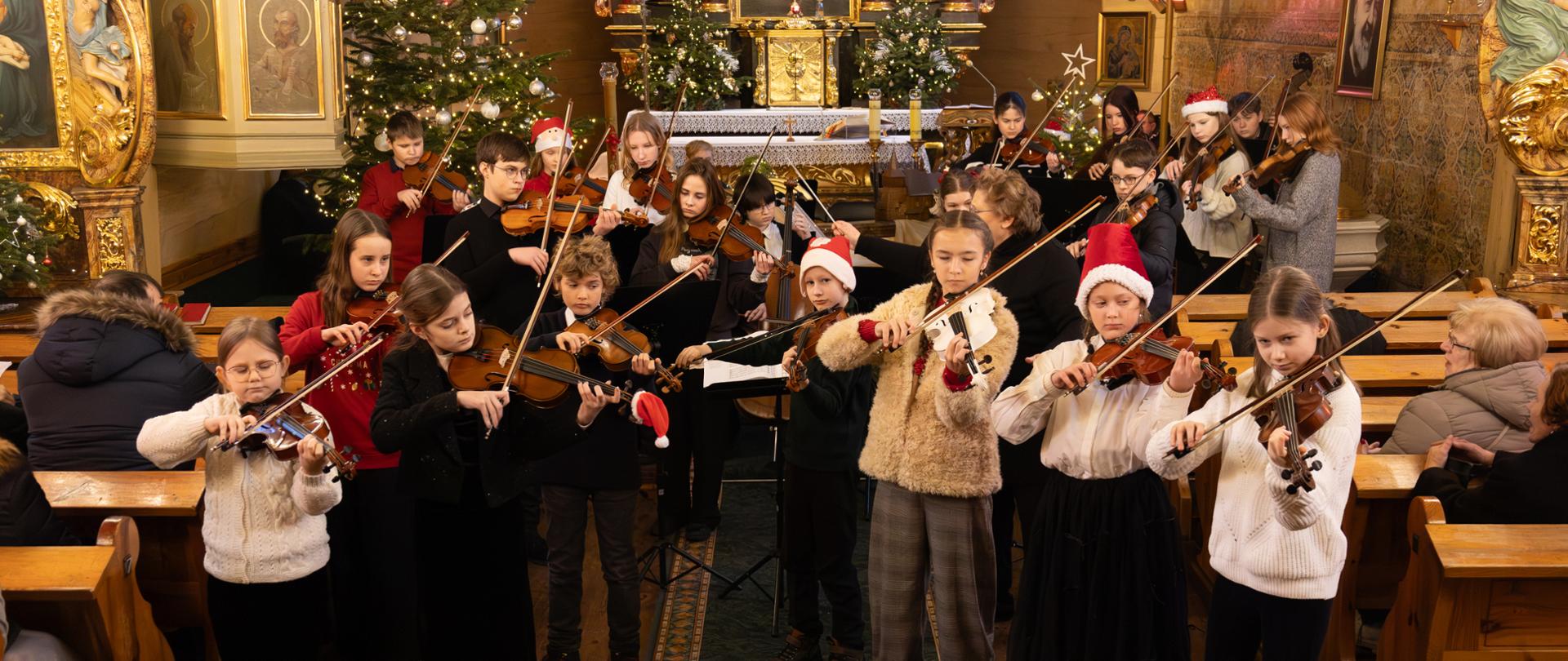 Na zdjęciu widać grupę dzieci grających na skrzypcach w kościele. Uczniowie stoją w nawie głównej między ławkami, a za nimi widać ołtarz ozdobiony choinkami i świątecznymi dekoracjami. Część dzieci ma na głowach czerwone czapki Mikołaja. W tle siedzą dorośli, którzy słuchają koncertu. Cała scena ma ciepły, świąteczny nastrój, a wnętrze kościoła jest bogato zdobione złotymi elementami i obrazami.