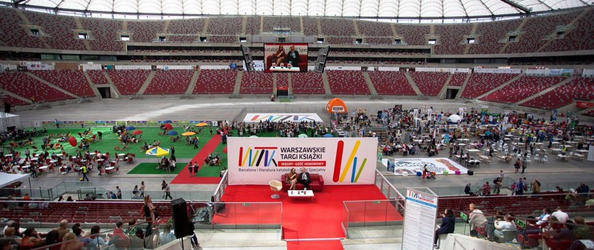 arena stadionu PGE Narodowy; na pierwszym planie duży plakat z napisem Warszawskie Targi Książki. Przejście do plakatu wyłożone czerwonym dywanem; przed plakatem stoi czerwona kanapa, na której siedzą i rozmawiają kobieta z mężczyzną; za plakatem arena stadionu wypełniona ludźmi i stoiskami