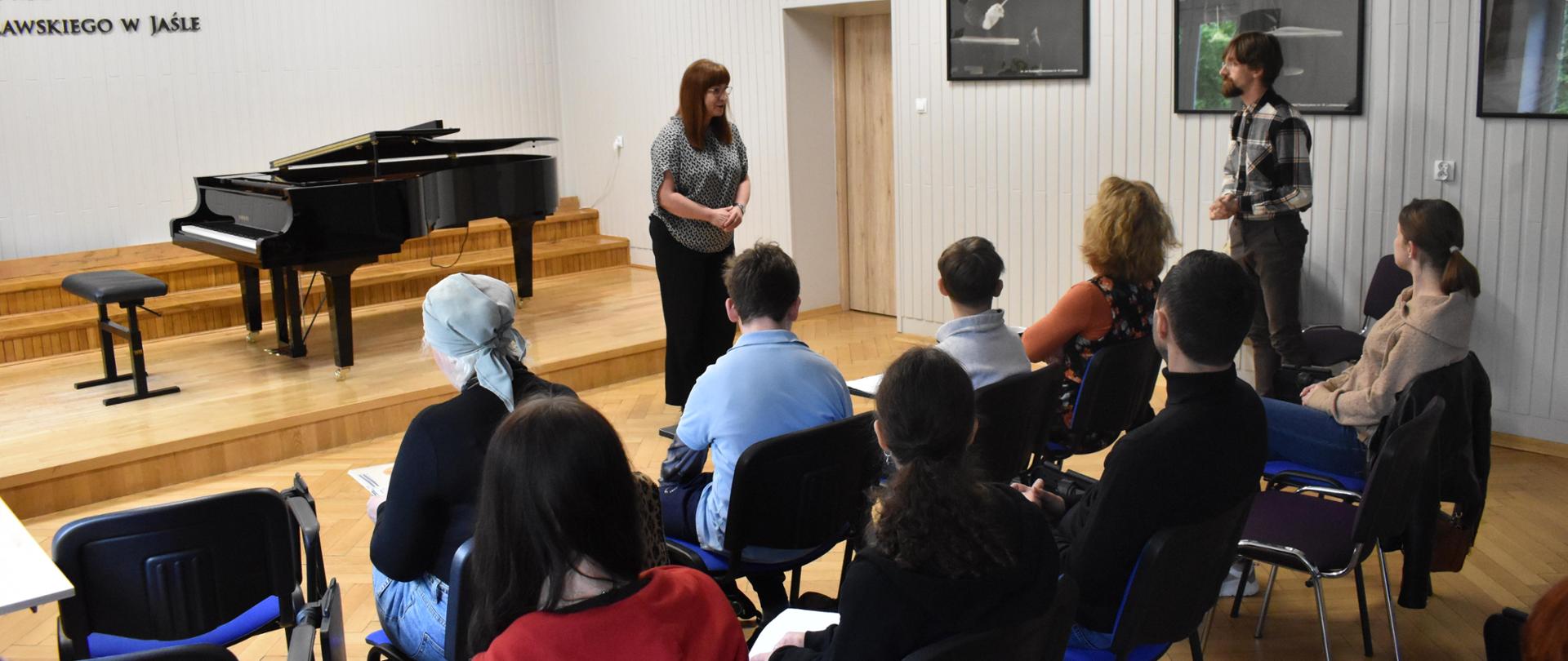 w auli PSM, podczas warsztatów pianistycznych widnieje p.dyrektor PSM I st. w Jaśle, która wita prowadzącego warsztaty pianistę oraz zgromadzoną społeczność szkolną. Kolorystyka zdjęcia jest biało-szaro-niebiesko-czerwono-czarna. 