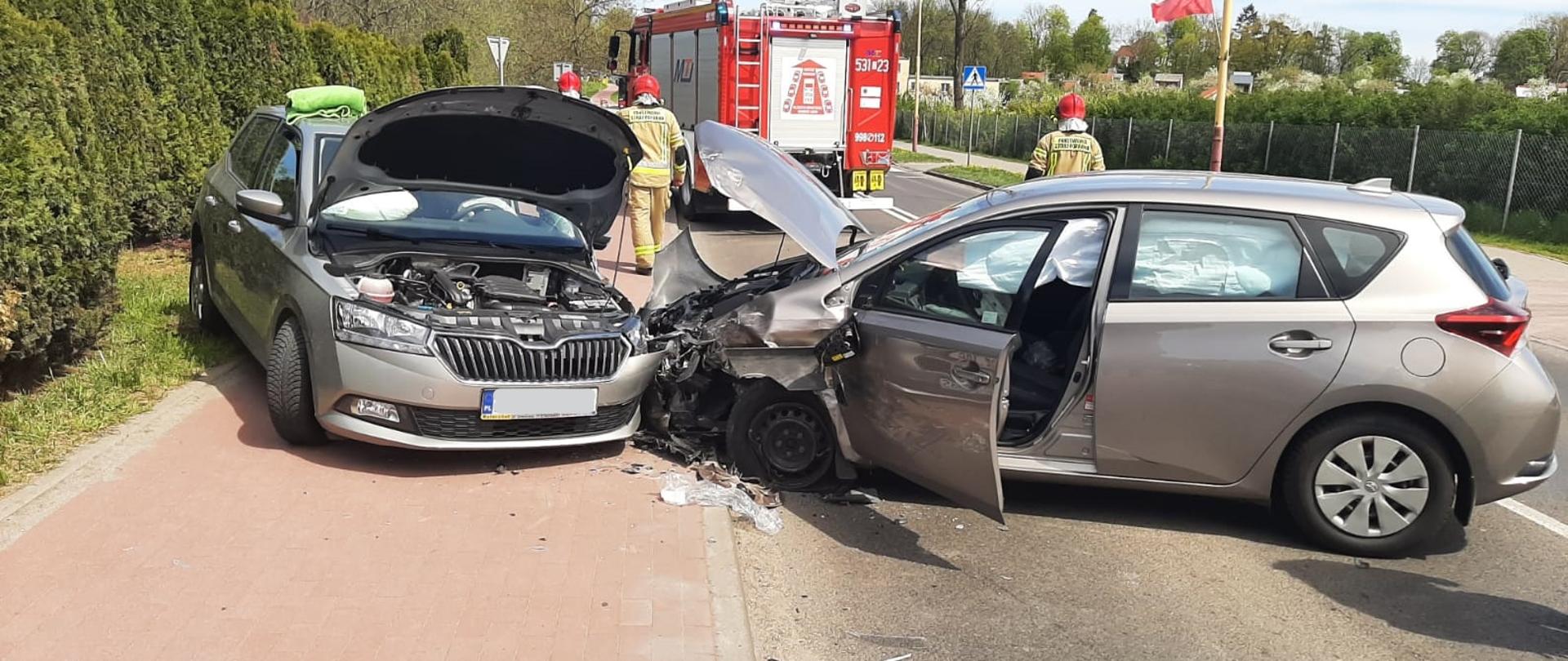 Uszkodzone pojazdy, w tle trzech strażaków i samochód pożarniczy