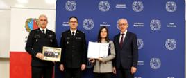 Zdjęcie pokazuje moment wręczenia dyplomu kobiecie – jednej z kursantek. Towarzyszą jej komendant główny PSP oraz pozostali przedstawiciele Akademii Pożarniczej. W rękach uczestniczki widoczny jest dyplom oraz medal.