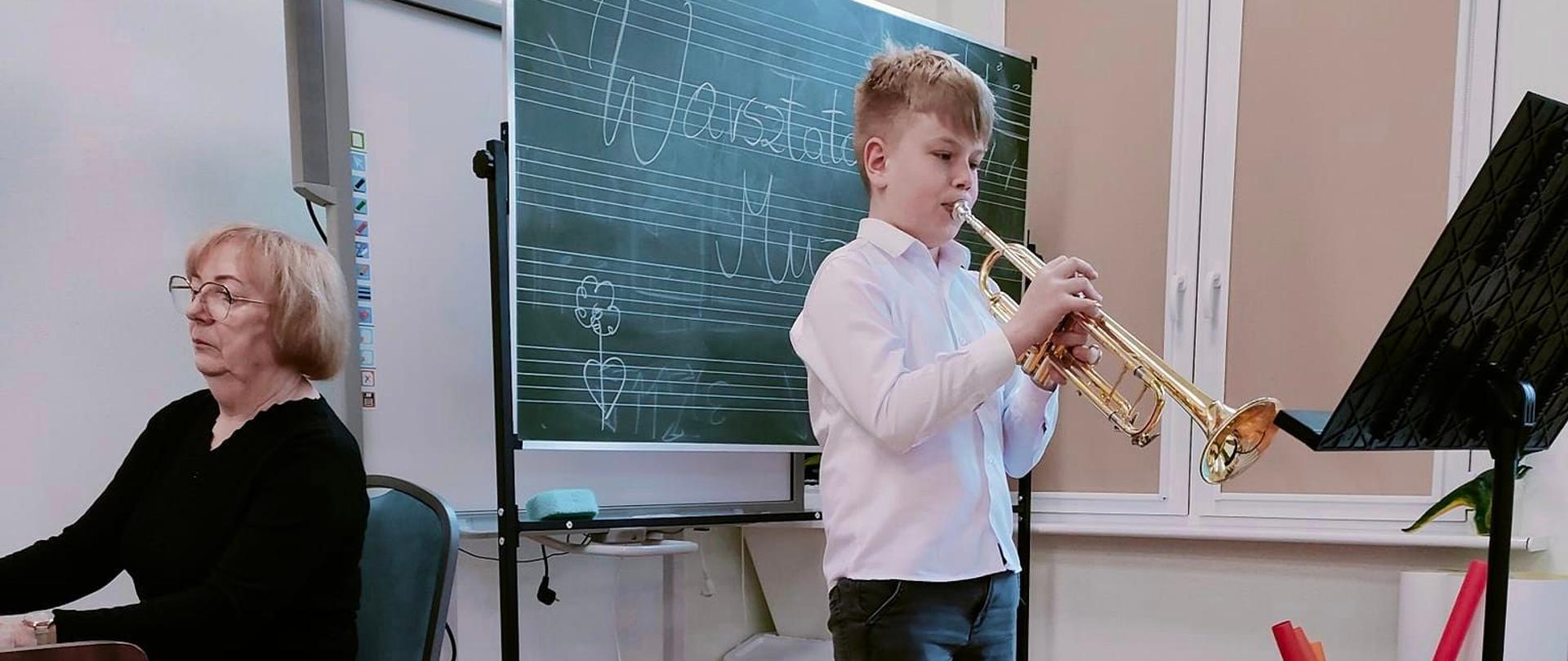 trębacz stojący w sali i grający audycję dla dzieci. Z lewej strony pani akompaniator przy fortepianie. 