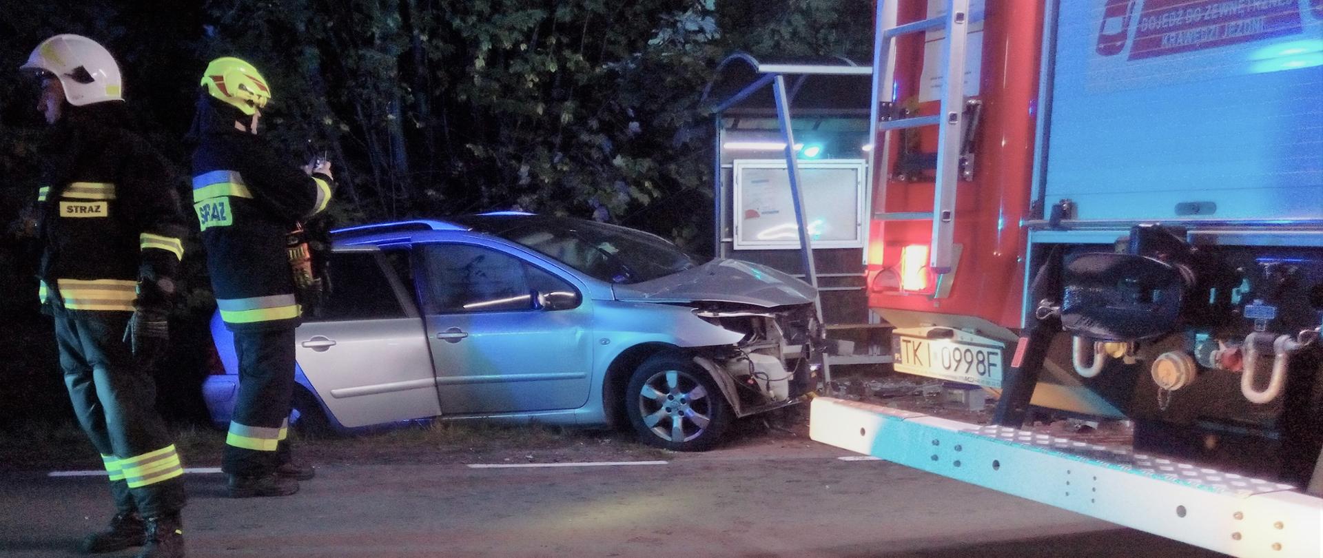 Zdjęcie przedstawia samochód osobowy i fragment wiaty przystankowej, w którą pojazd uderzył. Po lewej stoją dwaj strażacy a po prawej widać fragment samochodu gaśniczego.