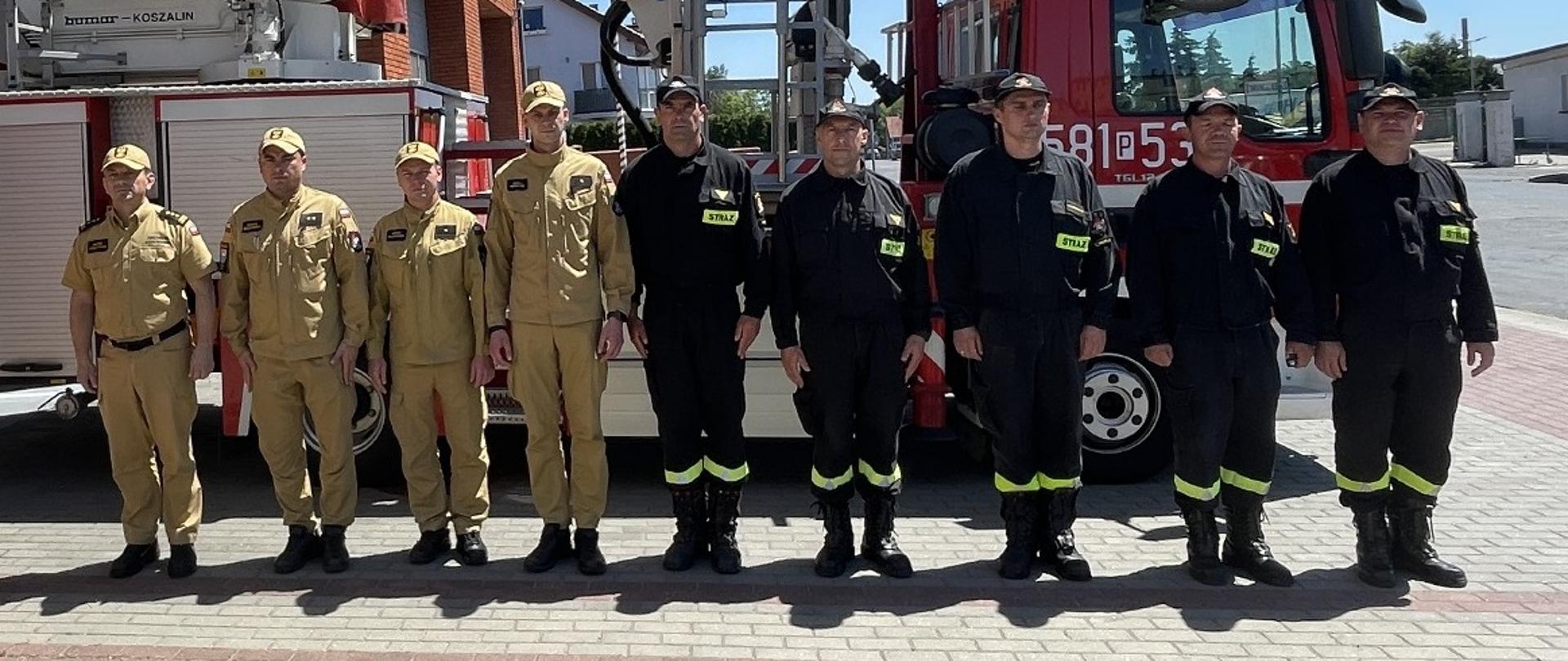 Strażacy stoją ustawienie w równym szeregu na tle wozu strażackiego - drabiny - na zewnątrz, przed bramą wjazdową do remizy.