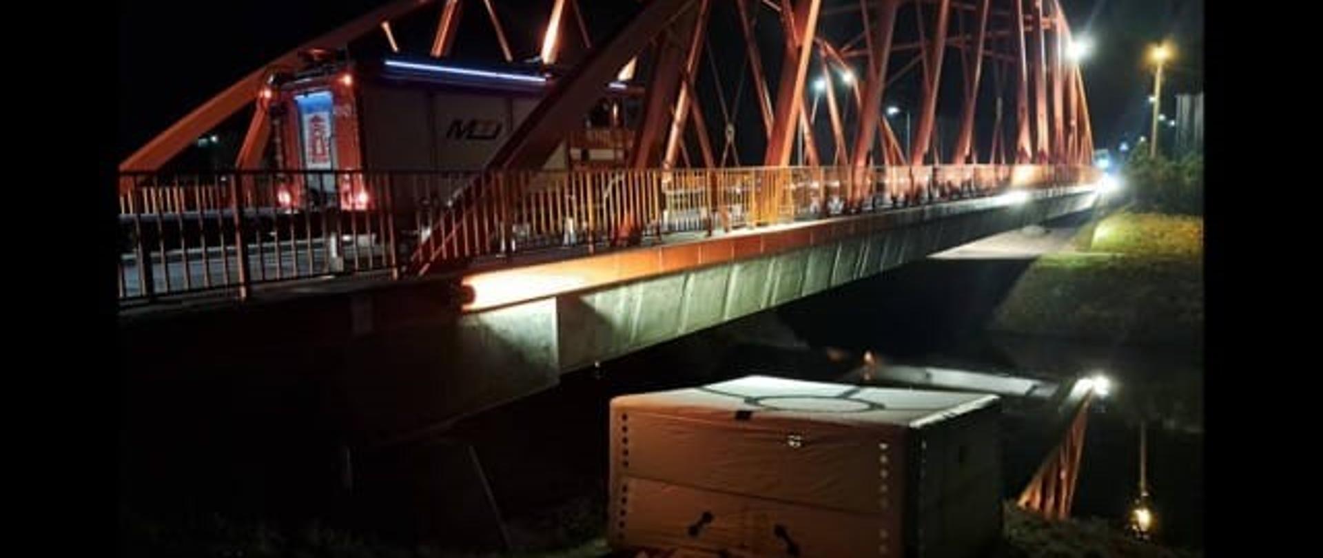 Zdjęcie zrobione nocą. Na zdjęciu oświetlony most z czerwonymi przęsłami. Na moście samochód strażacki. Pod mostem rozstawiony biały skokochron.