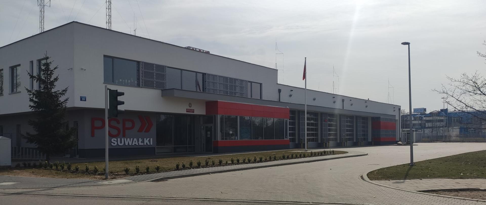 Na zdjęciu budynek komendy miejskiej państwowej straży pożarnej składający się z dwóch kondygnacji. w dalszej perspektywie garaże na pojazdy pożarnicze. Po lewej stronie budynku napis "PSP SUWAŁKI" obok wejście główne. Budynek w kolorze biało szarym z czeronymi wstawkami dookoła okien. Na budynku widać maszty antenowe. 