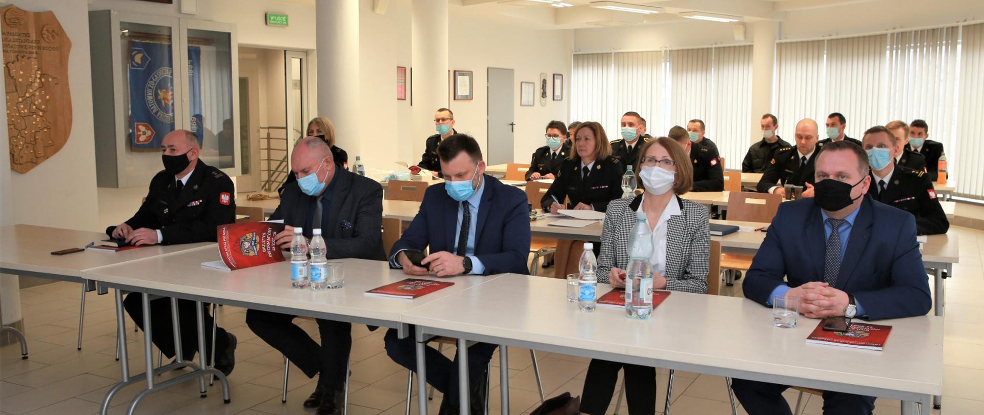 Zdjęcie w centralnej części przedstawia samorządowców lokalnych oraz Zastępcę komendanta powiatowego PSP w Bochni, którzy siedzą przy stoikach i słuchają wygłaszanej narady. Na stolikach leżą biuletyny KP PSP w Bochni. Na dalszym planie widać strażaków, którzy również biorą udział w naradzie