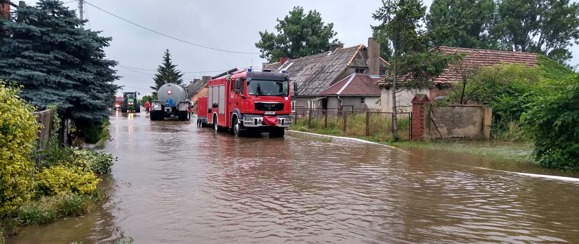 Zalana jezdnia w tle gospodarstwa a na jezdni samochód pożarniczy oraz beczkowóz i ciągnik 