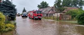 Zalana jezdnia w tle gospodarstwa a na jezdni samochód pożarniczy oraz beczkowóz i ciągnik 