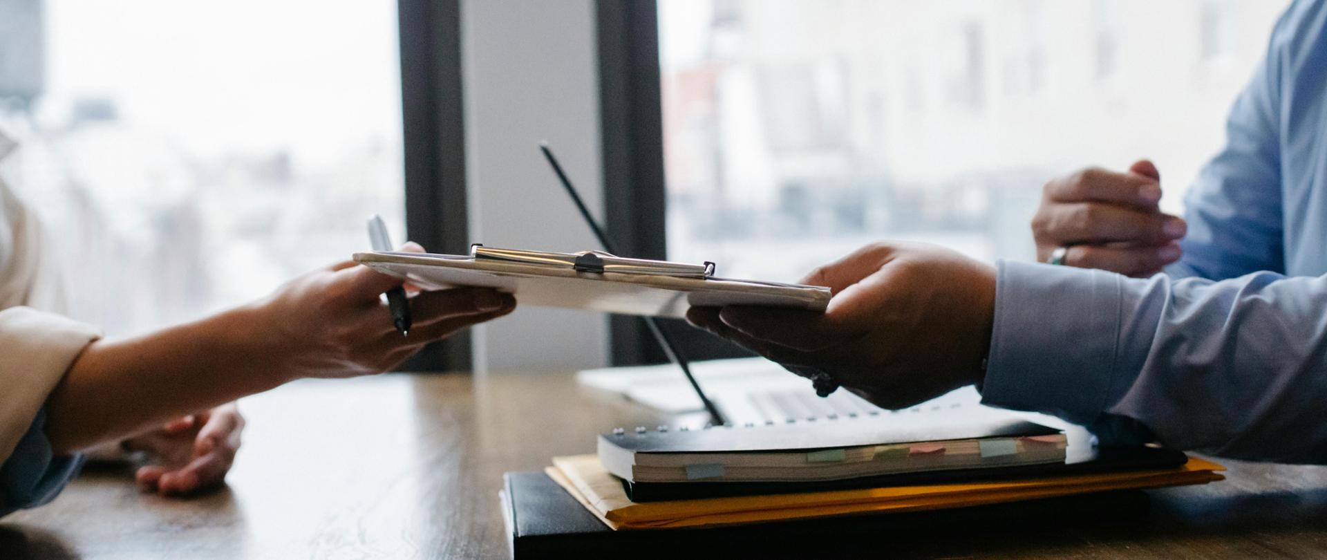 Dwójka ludzi przekazuje sobie dokument nad stołem na którym leżą zeszyty, teczki i listy