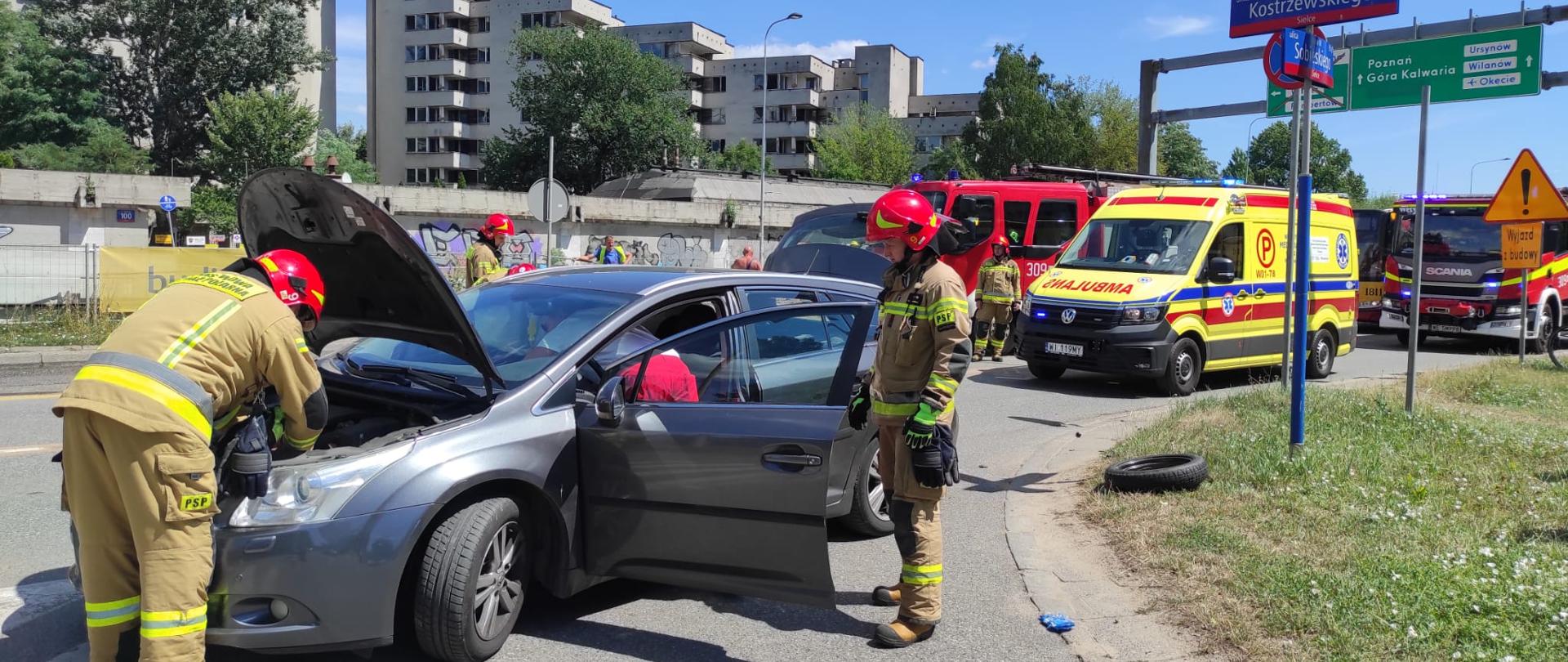 Na zdjęciu widać pracujących strażaków, samochód osobowy z otwartą maską, który brał udział w zdarzeniu. Na zdjęciu widoczna jest karetka pogotowia oraz wozy straży pożarnej. 