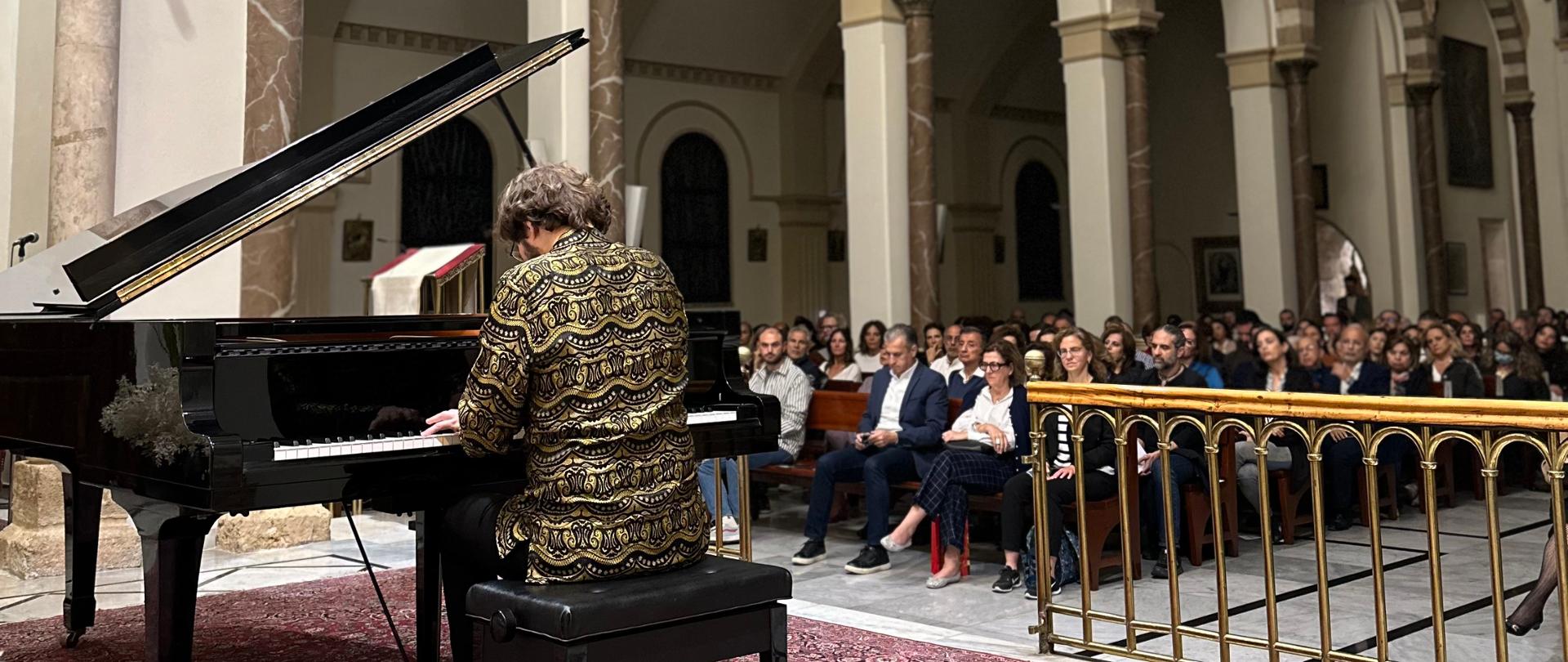 Pianista gra na fortepianie, a w tle publiczność siedzi w ławkach kościoła i słucha koncertu 