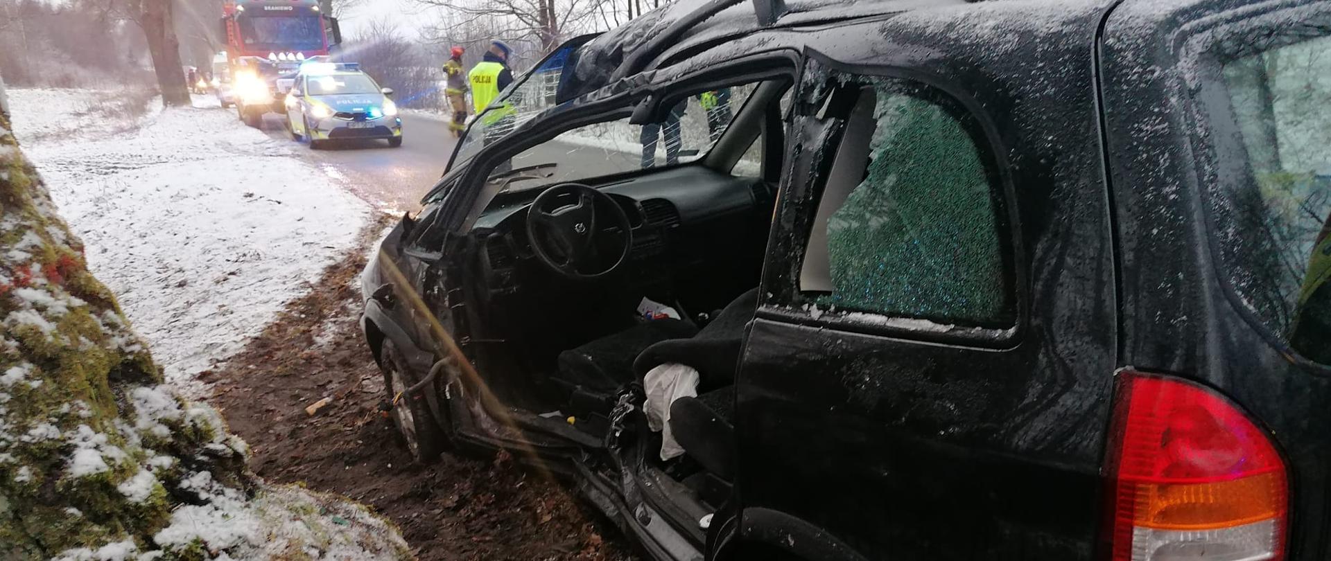 Uszkodzone ciemne auto po uderzeniu w drzewo. W oddali pojazdy ratownicze.