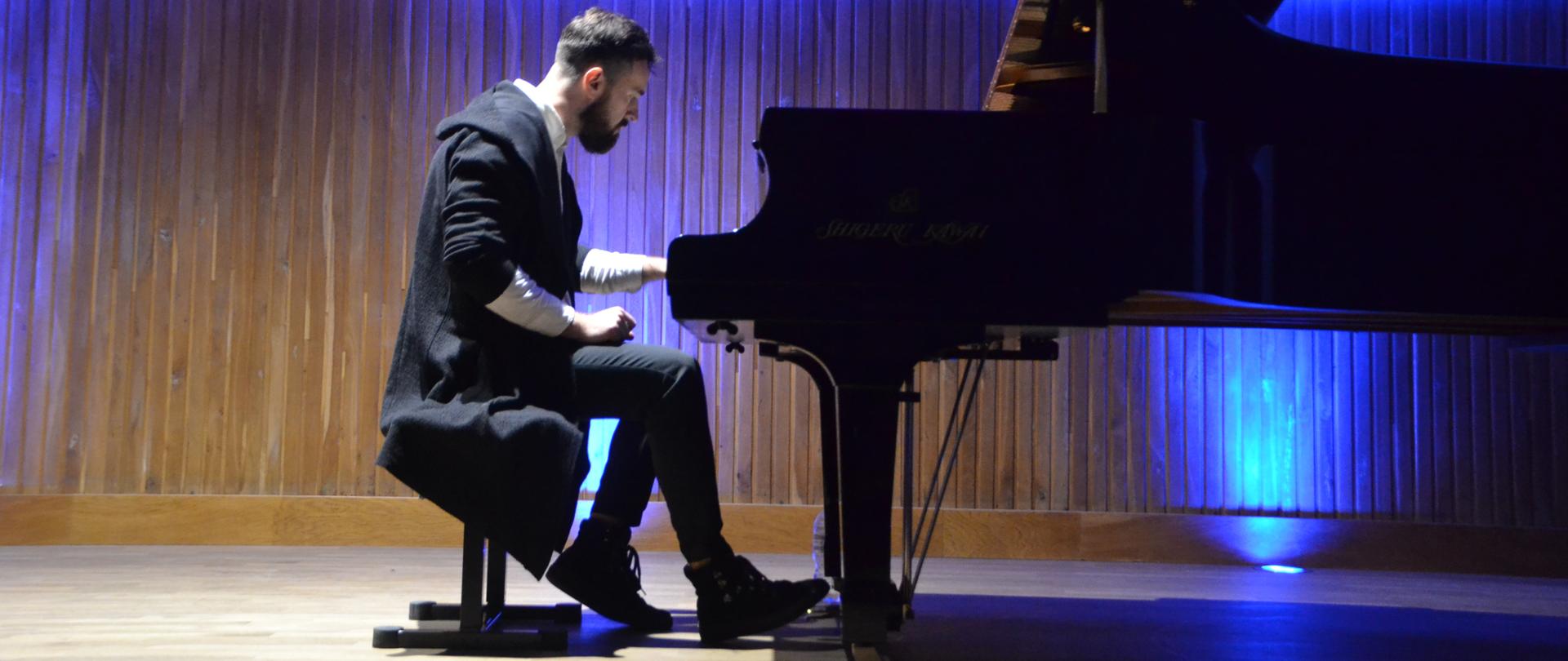 Zdjęcie w części centralnej przedstawia pianistę grającego na fortepianie we wnętrzu sali koncertowej. Tło zdjęcia jest rozświetlone lampami emitującymi kolor niebieski.