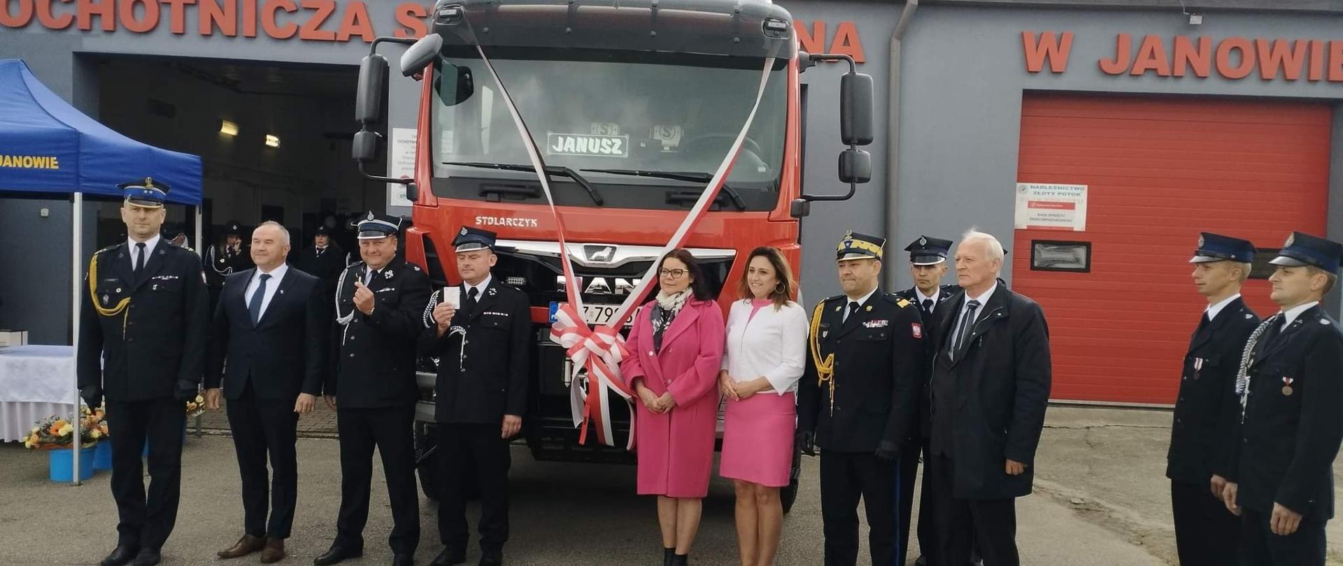 Na zdjęciu widać funkcjonariuszy przed remizą OSP Janów podczas uroczystego przekazania pojazdu. Na pojeździe powieszona wstęga. W tle widoczny budynek remizy. Na pierwszym planie funkcjonariusze Państwowej i Ochotniczej Straży Pożarnej oraz zaproszeni goście.