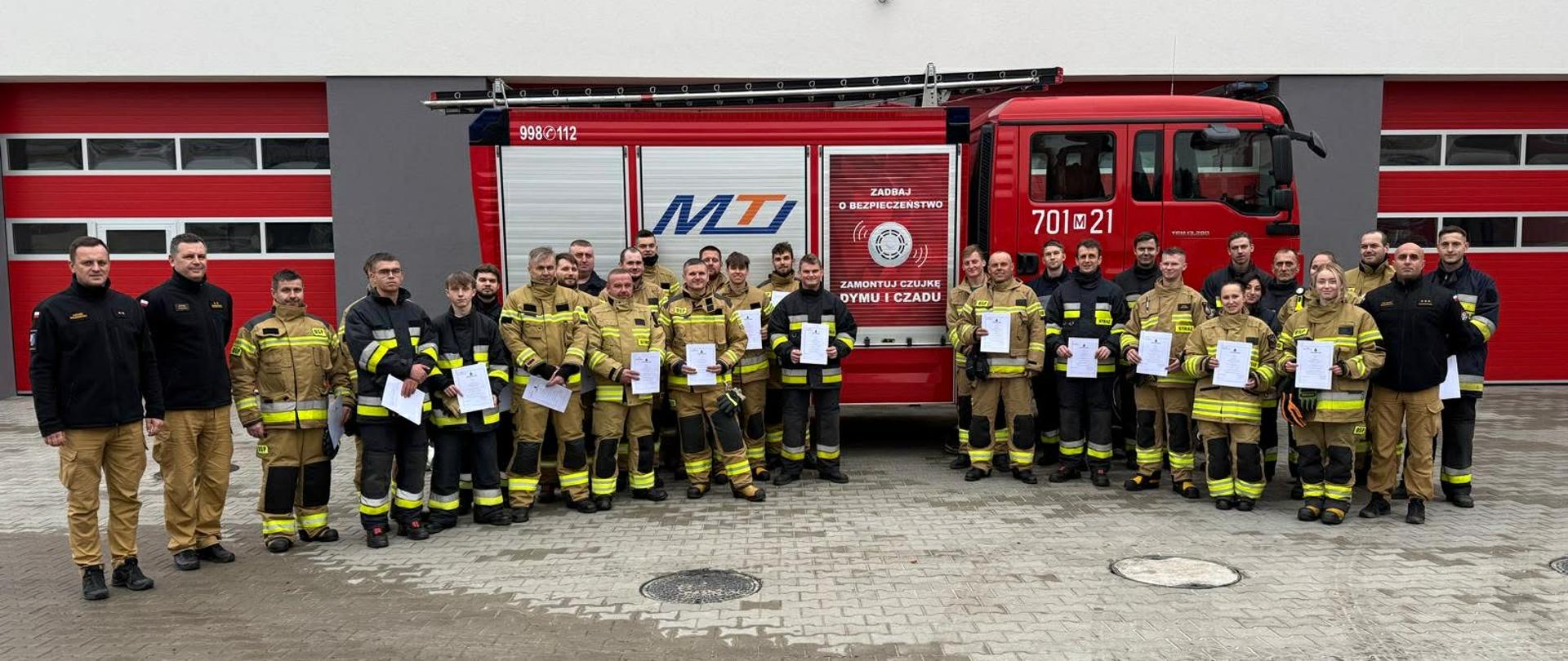 Duża grupa strażaków pozuje do zdjęcia przed czerwonym wozem strażackim i budynkiem strażnicy. Każda osoba trzyma w rękach dokument, prawdopodobnie certyfikat lub zaświadczenie ukończenia szkolenia.
