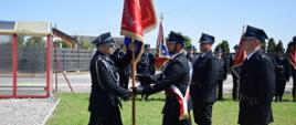 Dwóch mężczyzn w mundurach strażackich przekazuje sobie sztandar. Obok nich stoją dwaj inni strażacy. W tle widać innych umundurowanych strażaków ze sztandarami, a także elementy zabudowań, ogrodzenia i fragment wiaty przystankowej. 