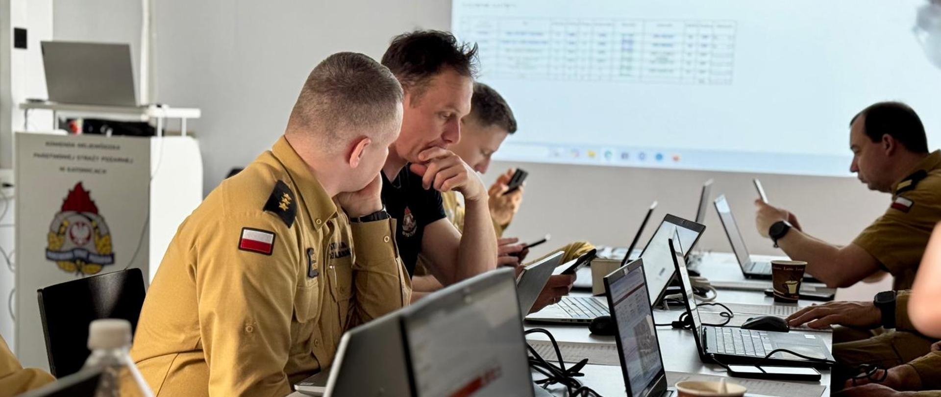 Zdjęcie przedstawia grupę strażaków w mundurach siedzących przy stole, na którym znajdują się laptopy i inne urządzenia elektroniczne. Osoby te są zaangażowane w pracę lub dyskusję, patrząc na ekrany swoich laptopów. W tle znajduje się ekran projekcyjny z wyświetlonymi danymi tabelarycznymi i wykresami. Na stole widoczne są również filiżanki z kawą oraz butelka wody. 