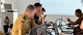 Zdjęcie przedstawia grupę strażaków w mundurach siedzących przy stole, na którym znajdują się laptopy i inne urządzenia elektroniczne. Osoby te są zaangażowane w pracę lub dyskusję, patrząc na ekrany swoich laptopów. W tle znajduje się ekran projekcyjny z wyświetlonymi danymi tabelarycznymi i wykresami. Na stole widoczne są również filiżanki z kawą oraz butelka wody. 