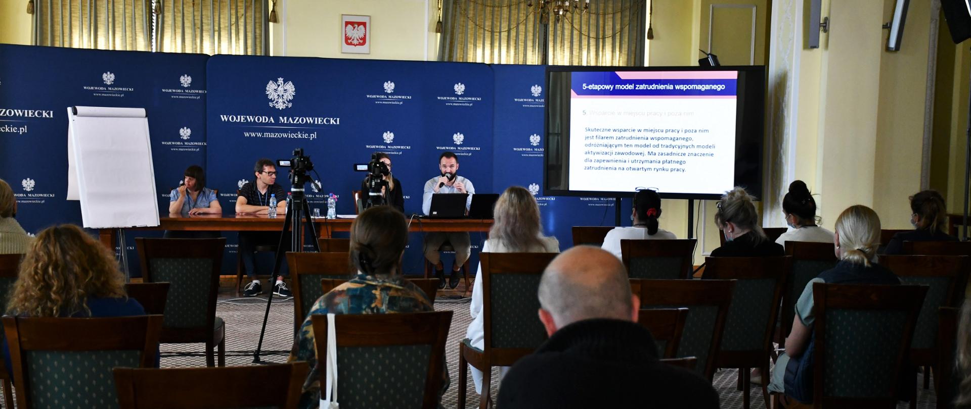 Na pierwszym planie zdjęcia znajduje się skromna widownia złożona z osób uczestniczących w wykładzie otwartym z zakresu zatrudnienia wspomaganego i współpracy z osobami z niepełnosprawnością. W tle znajduje się mały telebim, na którym odtwarzana jest prezentacja. Tuż obok telebimu znajduje się stół prezydialny, za którym zasiadło czworo prelegentów. Tuż za prelegentami znajdują się granatowe ścianki wizerunkowe Wojewody Mazowieckiego. 