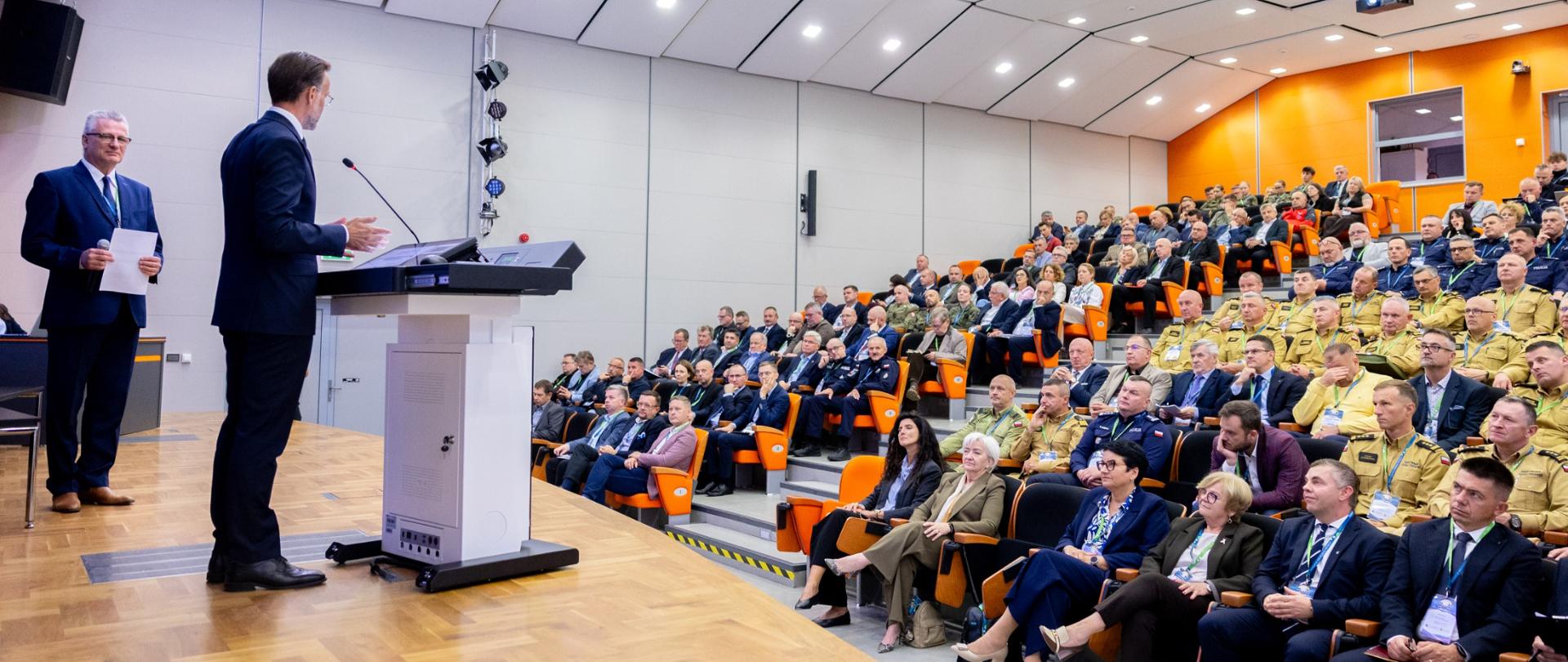Sala konferencyjna, widać zgromadzonych uczestników i mówcę na scenie.