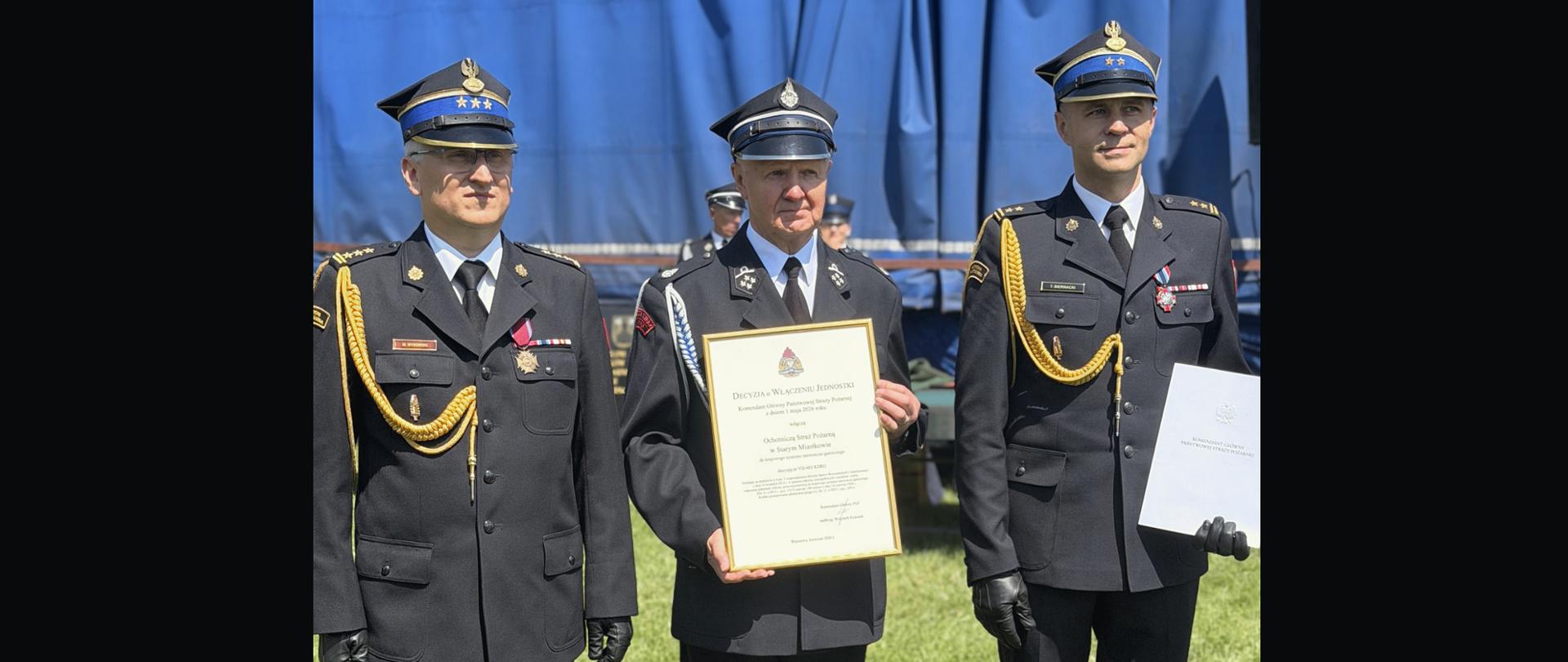 Trzej strażacy w galowych mundurach pozują do zdjęcia na tle niebieskiego namiotu. Środkowy z nich z dumą prezentuje oprawiony w ramę oficjalny dyplom uznania.
