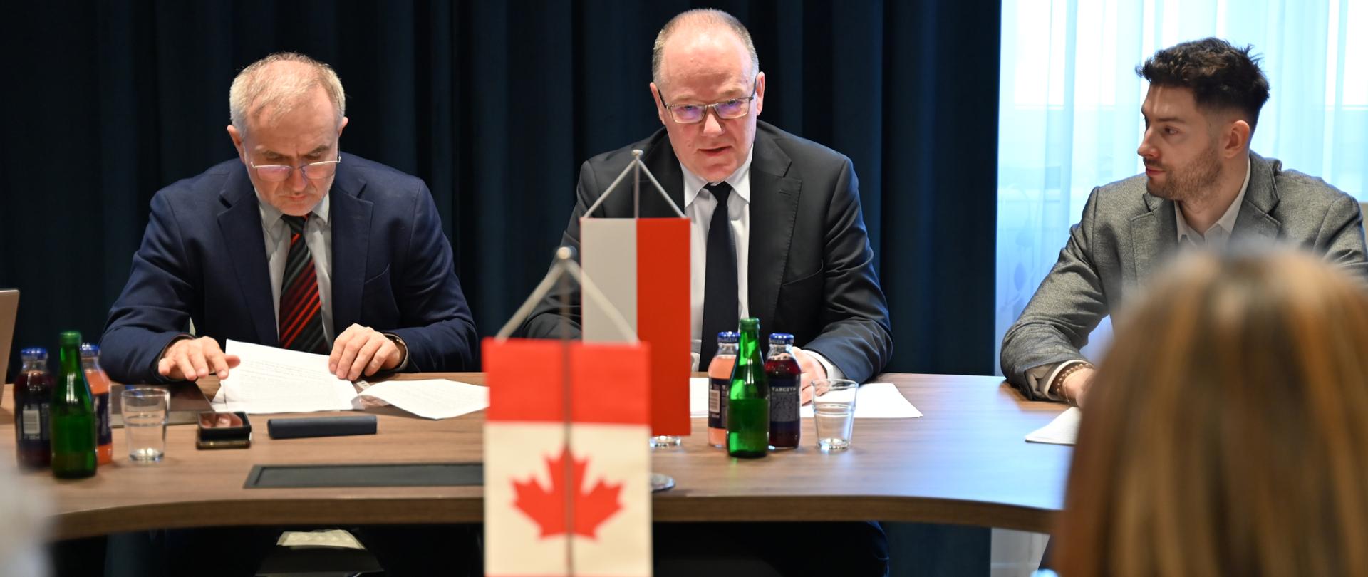 Wiceminister Krzysztof Galos, Główny Geolog Kraju, spotkał się z ambasador Kanady Catherine Godin, Na pierwszym planie widać mężczyznę przy stole mówiącego do siedzącej tyłem do widza kobiety. Mężczyzna siedzi za małą flagą Polski ustawioną na stole. Przed kobietą, której widać tylko włosy i plecy widać flagę kanady. Po obu stronach mężczyzny siedzi dwóch mężczyzn - ekspertów. 