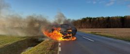 Pożar samochodu. Droga wojewódzka nr 434 w okolicach miejscowości Roszkówko. Na tle pola i lasu pali się ciemny samochód osobowy stojący na jezdni. Płomienie wydobywają się z przodu i tyłu auta.Towarzyszy temu czarny dym.