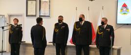 Funkcjonariusz Państwowej Straży Pożarnej melduje kierownictwu KW PSP rozpoczęcie uroczystości. Obok stoją dodatkowo dwaj strażacy. funkcjonariusze ubrani w mundury galowe.