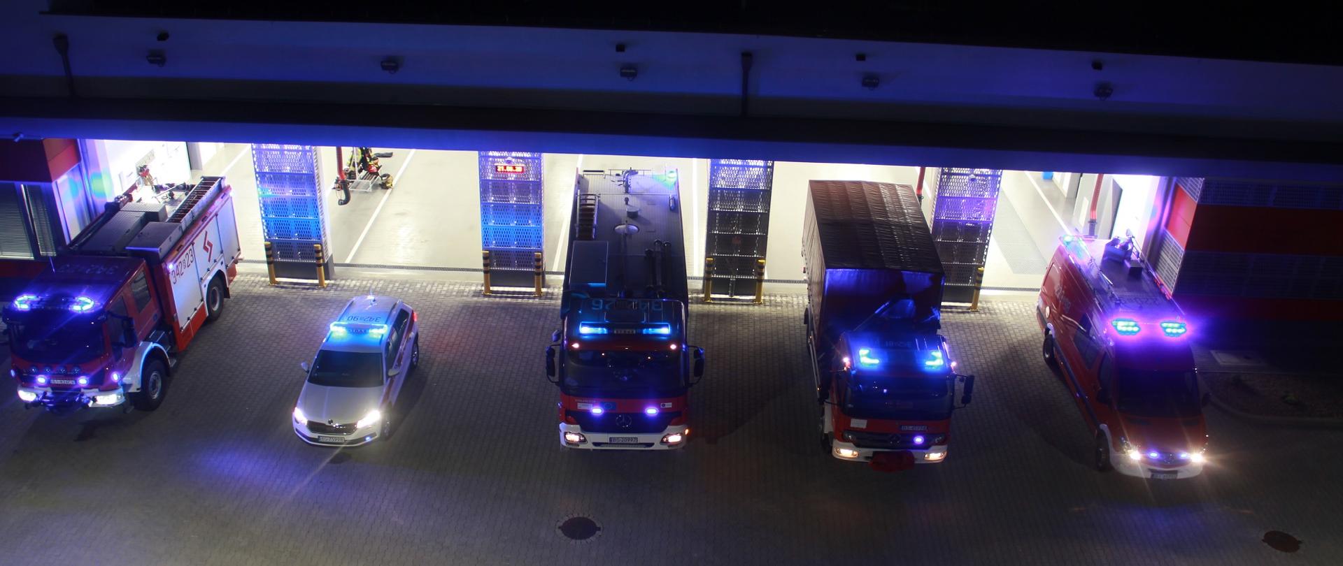 Na zdjęciu widać w nocy przed bramami garaży 5 samochodów strażackich z włączonymi sygnałami świetlnymi.
