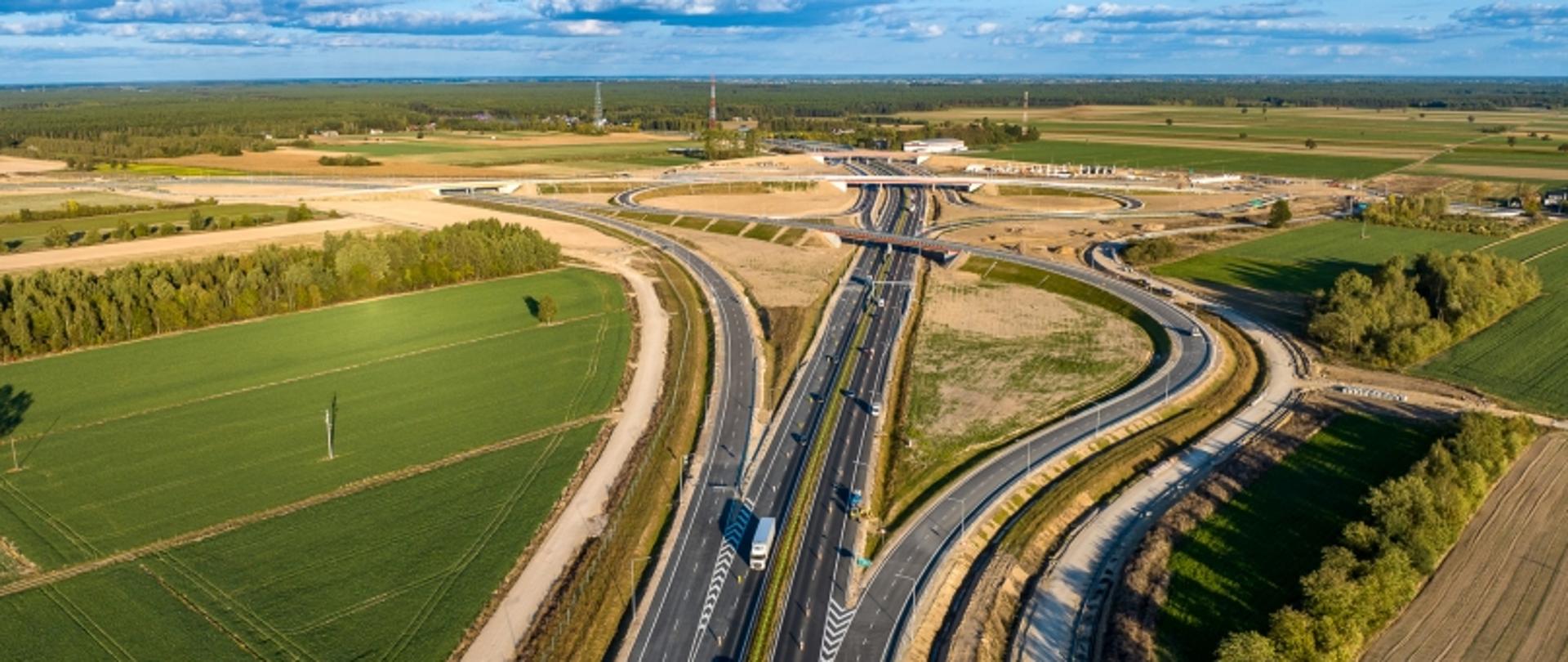 Aerial view na węzeł autostradowy otoczony polami i lasami, z estakadami i zakrętami.