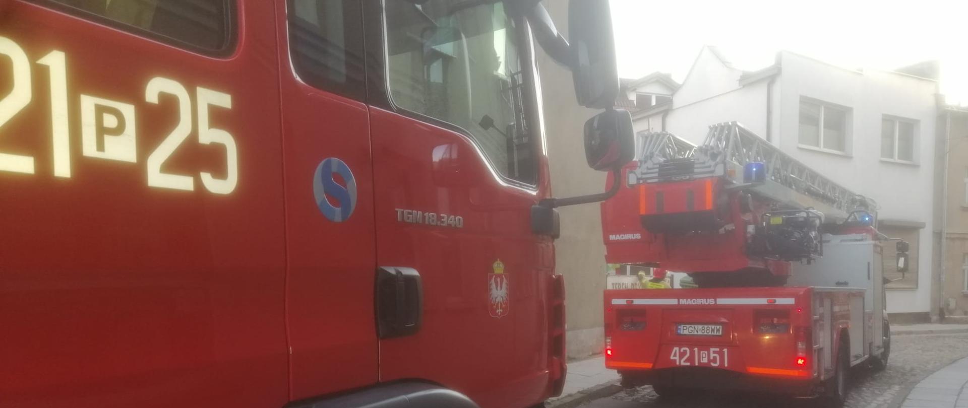 Na zdjęciu widać dwa samochody pożarnicze zadysponowane do pożaru mieszkania, które stoją na drodze jednokierunkowej