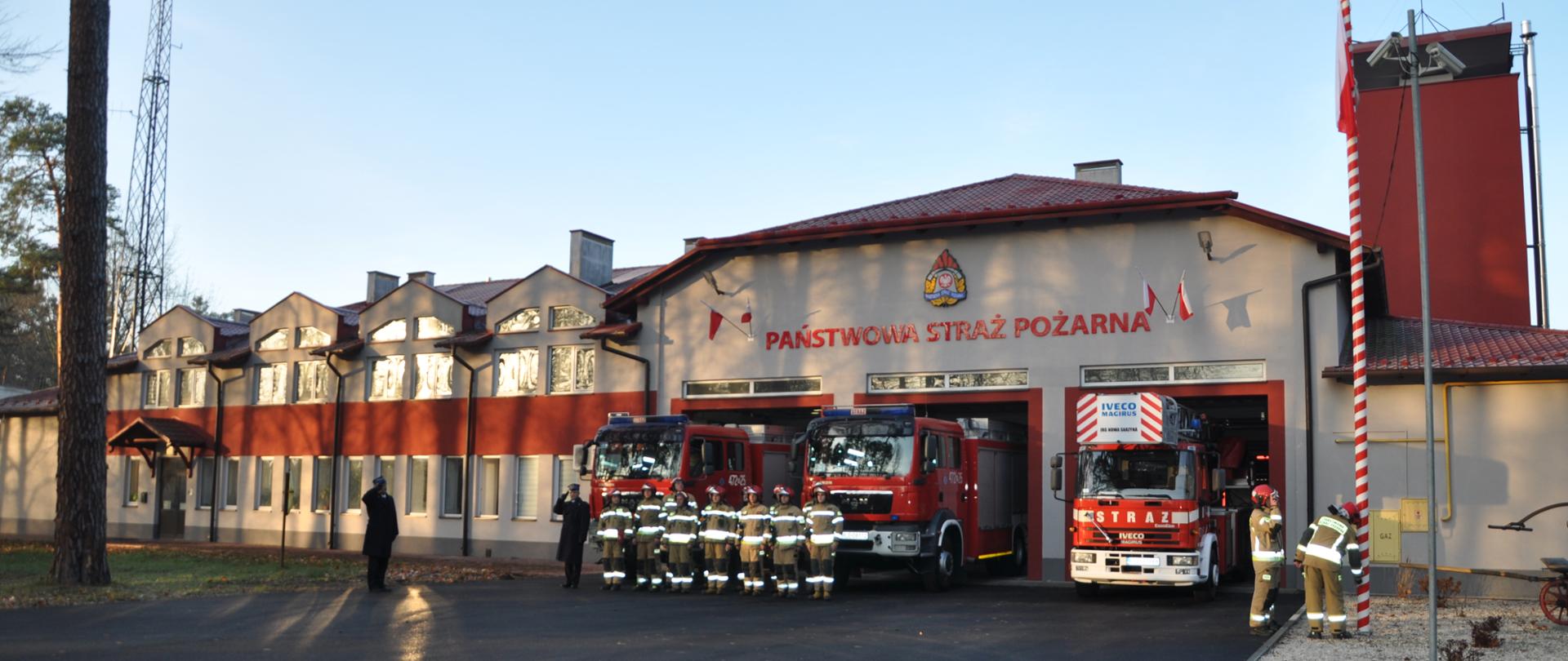 Na zdjęciu widzimy strażaków w pełnym umundurowaniu, którzy zebrali się aby uczcić 103 rocznicę odzyskania przez Polskę niepodległości. Stoją na baczność na tle budynku Jednostki Ratowniczo Gaśniczej.