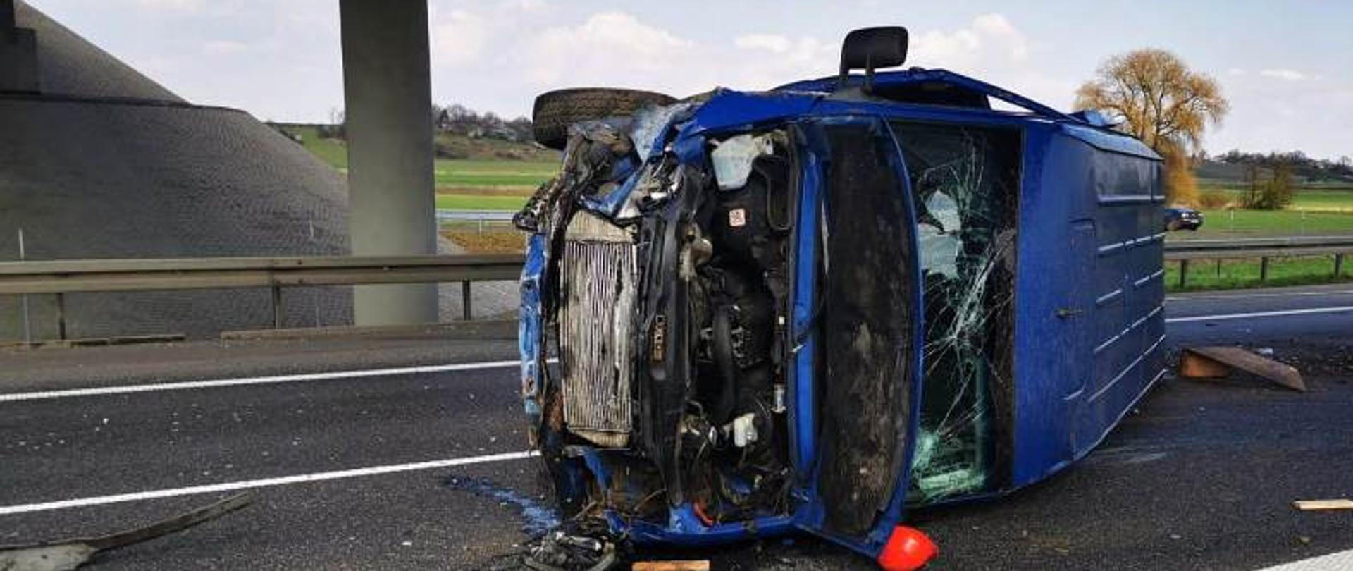 Zdjęcie przedstawia niebieski samochód dostawczy po wypadku drogowym. Samochód leży na lewym boku na pasie drogowym.