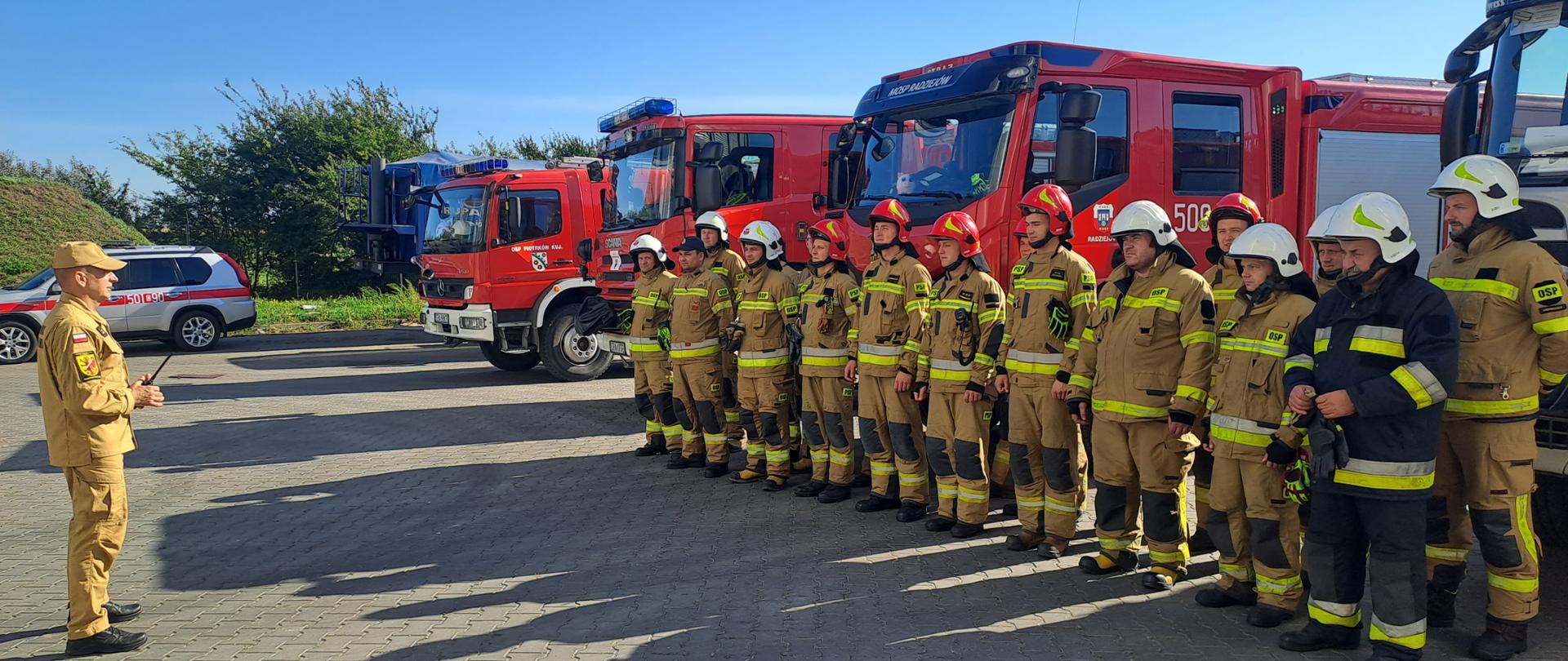 Zdjęcie przedstawia strażaków PSP oraz OSP podczas odprawy przed rozpoczęciem ćwiczeń ma drugim planie wozy straży pożarnych.