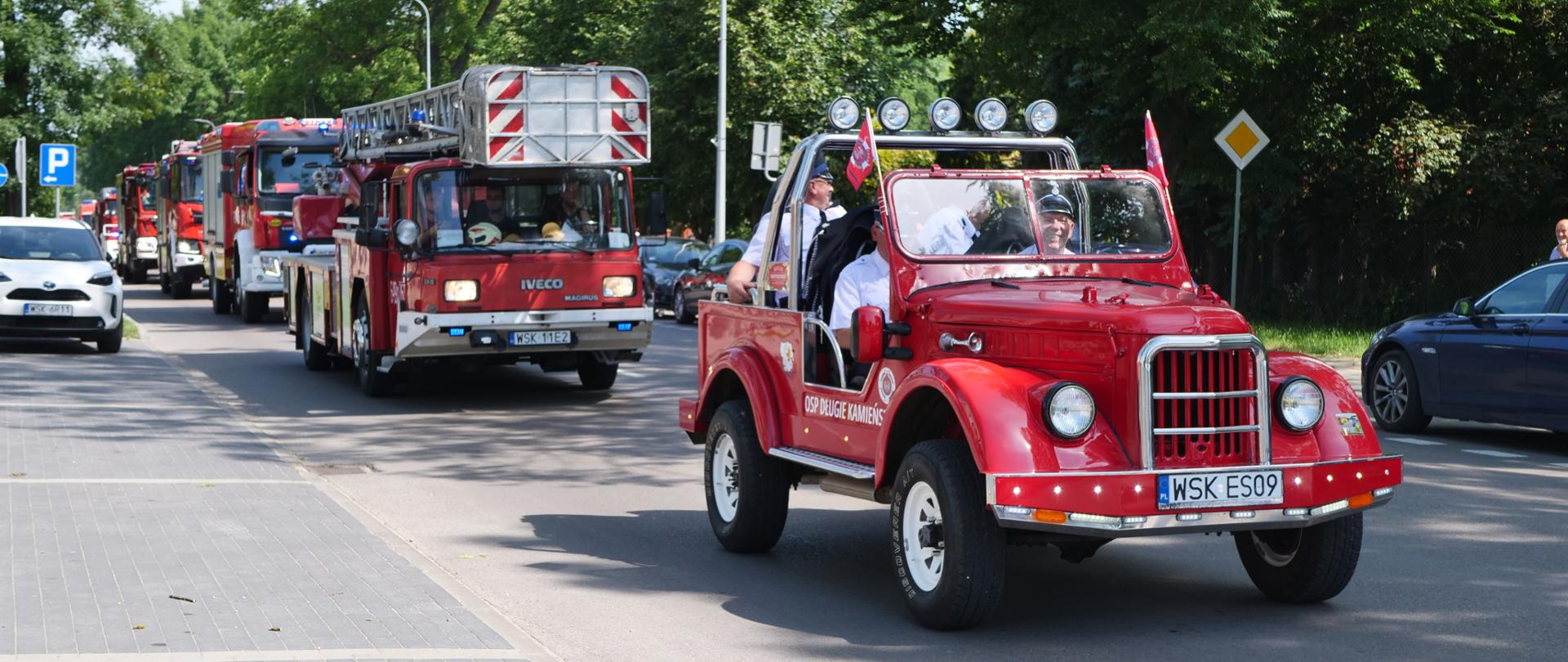 Zdjęcie przedstawia uroczystą paradę pojazdów pożarniczych poruszających się kolumną przez miasto. Na pierwszym planie jedzie czerwony, zabytkowy samochód gaśniczy bez dachu (typu retro/cabrio) z oznaczeniem „OSP DŁUGIE KAMIEŃSKIE”, w którym siedzą uśmiechnięci druhowie w białych koszulach. Za nim podąża nowoczesny wóz drabiniasty marki Iveco Magirus oraz inne współczesne samochody ratowniczo-gaśnicze na sygnale.