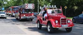 Zdjęcie przedstawia uroczystą paradę pojazdów pożarniczych poruszających się kolumną przez miasto. Na pierwszym planie jedzie czerwony, zabytkowy samochód gaśniczy bez dachu (typu retro/cabrio) z oznaczeniem „OSP DŁUGIE KAMIEŃSKIE”, w którym siedzą uśmiechnięci druhowie w białych koszulach. Za nim podąża nowoczesny wóz drabiniasty marki Iveco Magirus oraz inne współczesne samochody ratowniczo-gaśnicze na sygnale.