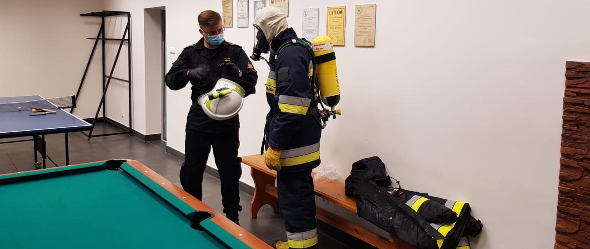Zdjęcie przedstawia funkcjonariusza Państwowej Straży Pożarnej. Strażak znajduje się w świetlicy w jednej ze strażnic jednostki Ochotniczej Straży Pożarnej. Ubrany jest w ubranie dowódczo - sztabowe koloru czarnego. Na twarzy ma założoną maseczkę ochronną. W lewej dłoni trzyma hełm bojowy. Na prawo od niego stoi strażak z Ochotniczej Straży Pożarnej. Ubrany jest w ubranie specjalne bojowe. Na głowie ma założoną kominiarkę i maskę podłączoną do aparatu powietrznego który ma na plecach. Funkcjonariusz Państwowej Straży Pożarnej prowadzi zajęcia z tematyki sprzęt ochrony dróg oddechowych. 