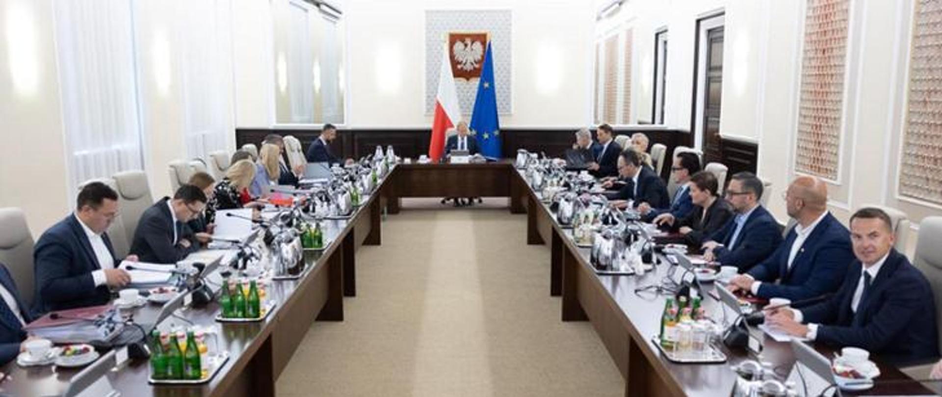 Trwają obrady dotyczące projektu ustawy. Przy stołach siedzą ministrowie uczestniczący w posiedzeniu. W tle widoczny jest premier, godło Rzeczypospolitej Polskiej oraz flagi Polski i Unii Europejskiej