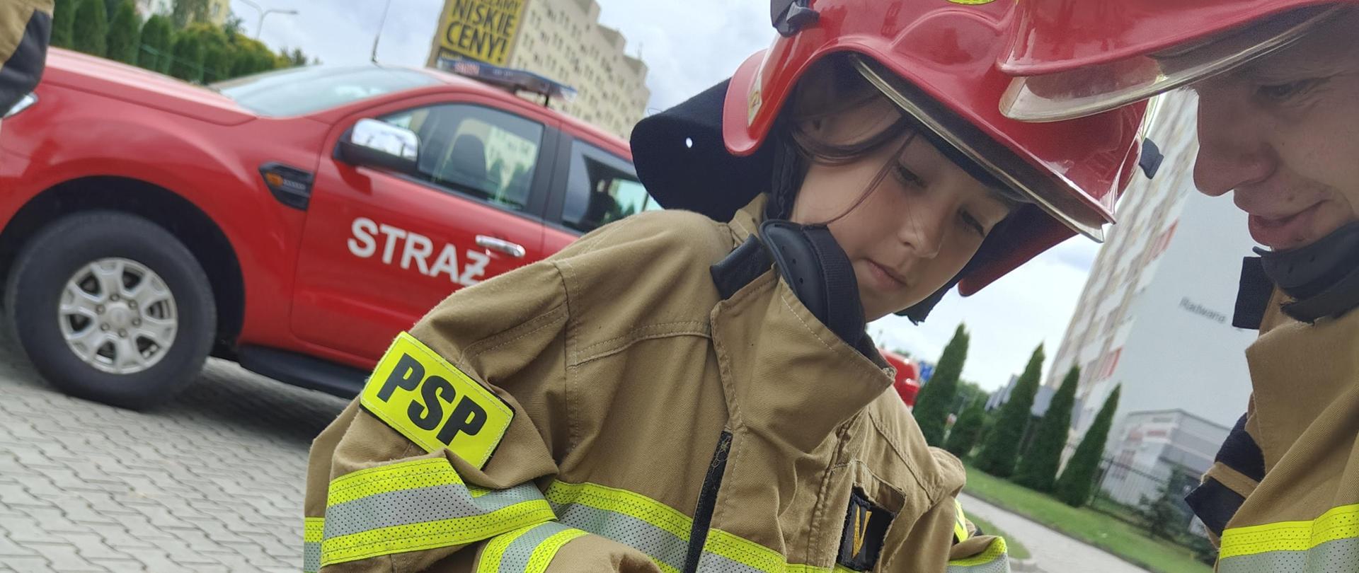 Dziewczynka podnosi nożyce hydrauliczne w asyście strażaka 