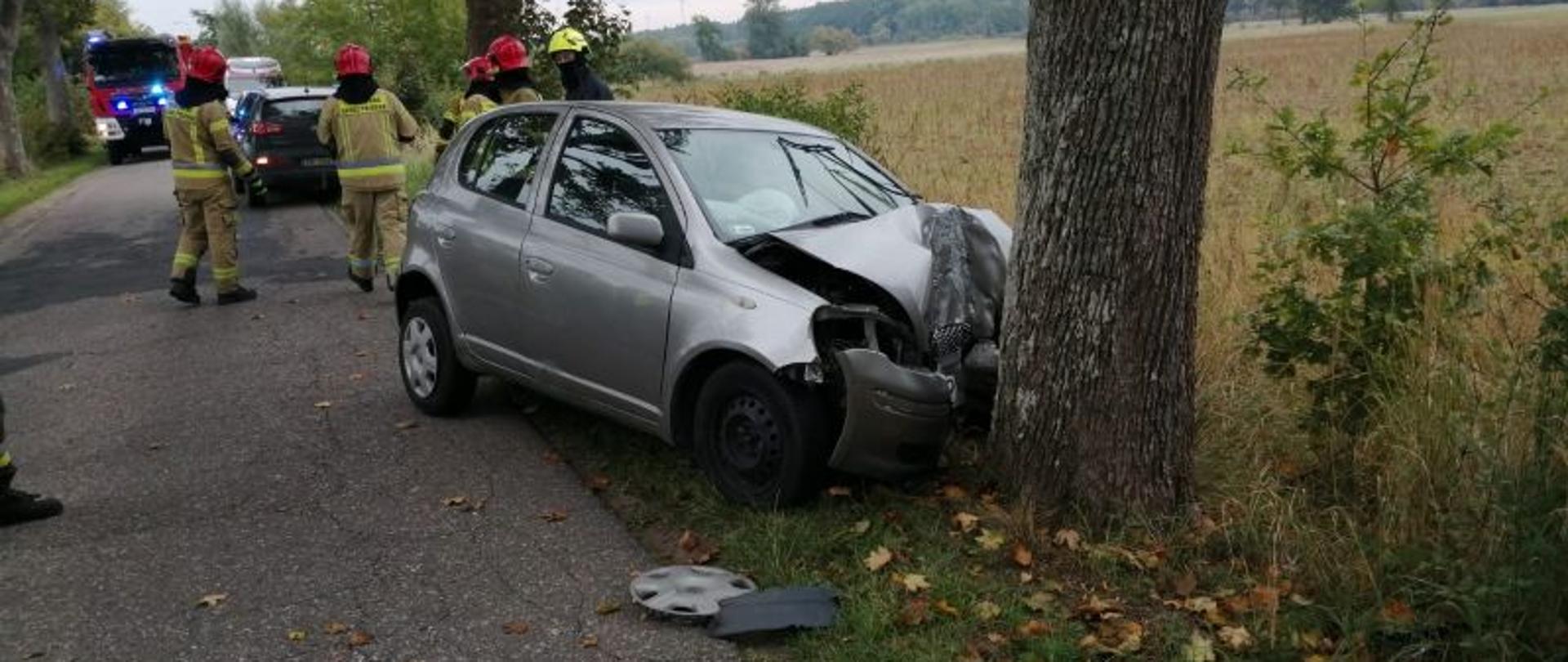 Zdjęcie przedstawia miejsce zdarzenia. Widać drogę, biegnące wzdłuż drogi drzewa. W jedno z drzew uderzyło auto osobowe. Małe, srebrne. Auto całkowicie zniszczone. Na miejscu widać działających strażaków. W oddali stoją auta ratunkowe.