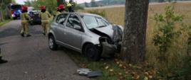 Zdjęcie przedstawia miejsce zdarzenia. Widać drogę, biegnące wzdłuż drogi drzewa. W jedno z drzew uderzyło auto osobowe. Małe, srebrne. Auto całkowicie zniszczone. Na miejscu widać działających strażaków. W oddali stoją auta ratunkowe.