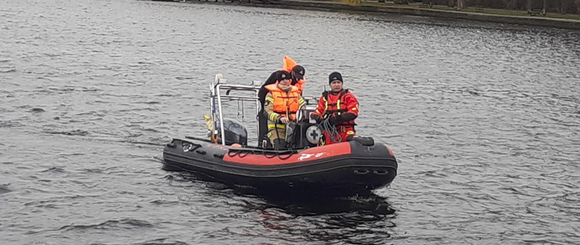 dwóch strażaków ubranych w ubranie specjalne i kamizelkę asekuracyjną wspólnie z przedstawicielem WOPR płyną pontonem ratowniczym WOPRU.