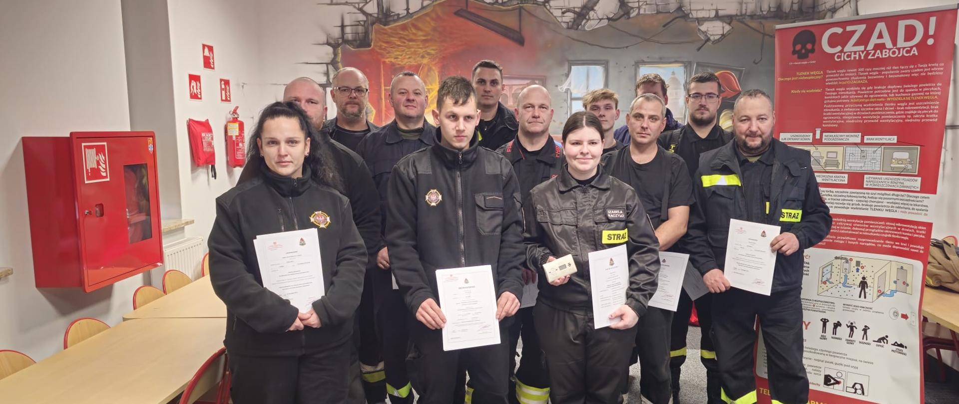 Zdjęcie przedstawia uczestników szkolenia podstawowego strażaka ratownika Ochotniczych Straży Pożarnych trzymających zaświadczenia ukończenia szkolenia (oraz w przypadku jednej druhny również czujki tlenku węgla) zlokalizowanych w sali edukacyjnej Komedy Powiatowej Państwowej Straży Pożarnej w Gołdapi